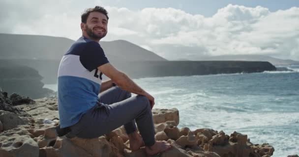 Joven chico atractivo con una gran sonrisa disfrutando del tiempo en medio de una increíble vista de la naturaleza con la montaña y el mar con grandes olas. 4k — Vídeo de stock