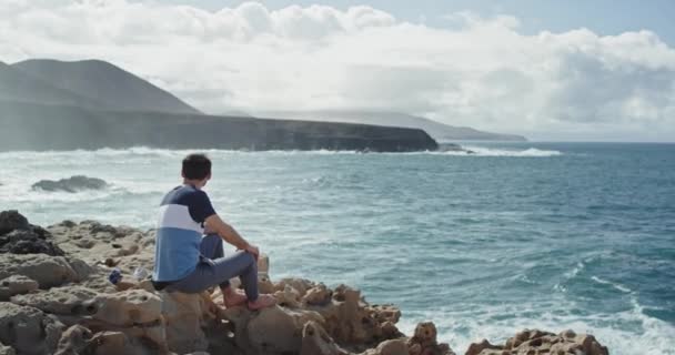 Fiatal srác a sziklákon élvezi a tájat. A tengerre és a hullámokra koncentrál. — Stock videók