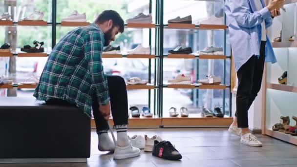 Em uma loja de sapatos um cliente cara tentar usar alguns sapatos novos outro cliente senhora tentar encontrar alguns sapatos das prateleiras — Vídeo de Stock