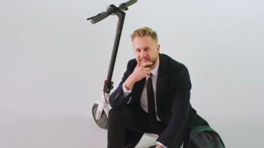 Stüdyoda kameranın önünde siyah takım elbiseli beyaz bir işadamı motosikletin yanında oturuyor ve mutlu bir şekilde kameraya bakıyor.