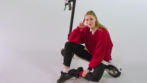 Sevimli bir gülümsemesi olan güzel bir bayan beyaz arka plandaki stüdyoda elektro scooterının yanında kameraya bakıyor. — Stok video