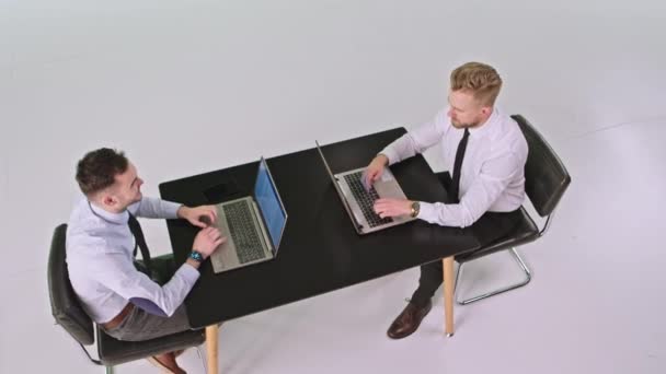 Studio d'arrière-plan blanc deux travailleurs hommes portant des vêtements de travail assis sur la table en utilisant deux ordinateurs portables pour travailler — Video