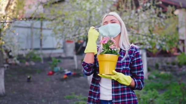 Femme aux cheveux blonds enlevant le masque de protection au milieu du jardin, elle tient le pot de fleurs et sourit grand coronavirus pandémique — Video