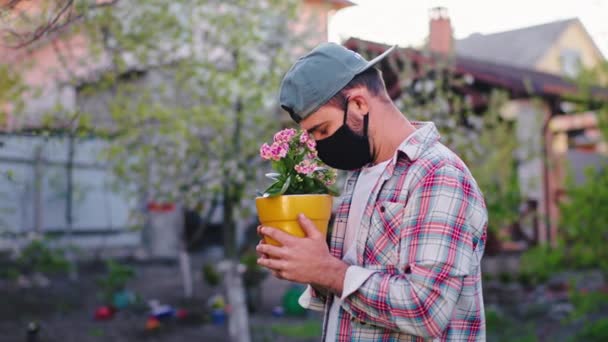L'uomo con una maschera protettiva che tiene il vaso di fiori in giardino a casa in pandemia di Coronavirus 2019 abbraccia con attenzione il vaso di fiori molto sconvolto. Girato su ARRI Alexa Mini — Video Stock