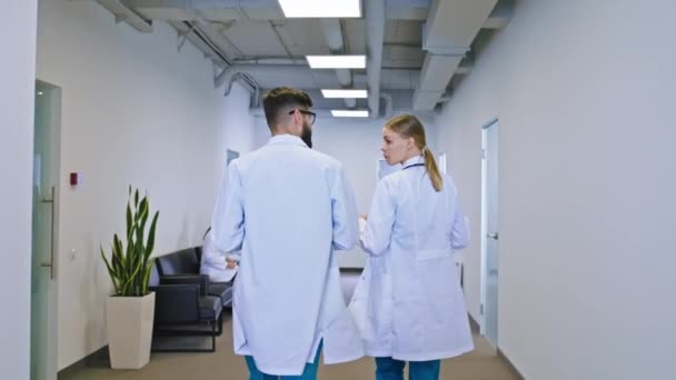 Δύο νεαρές νοσοκόμες κυρία και ο τύπος περπατώντας μέσα από το διάδρομο του νοσοκομείου και συζητώντας κάποια προβλήματα κρατώντας το χάρτη με σημειώσεις άλλοι γιατροί κάθονται στην καρέκλα — Αρχείο Βίντεο