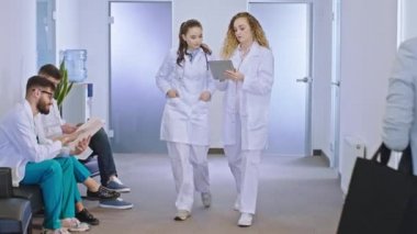 Modern hastane koridorunda, ana doktor kadın, meslektaşına kameranın önünde yürürken hastanın teşhisini dijital tabletle açıklıyor.