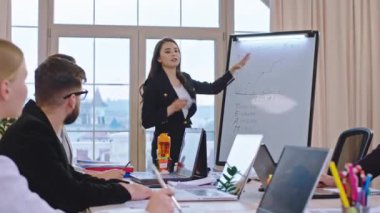 Genç bir kadın beyaz tahtada yeni bir iş planını ve istatistikleri açıklıyor. Birlikte analiz ediyorlar ve bazı liderlik fikirleri veriyorlar.