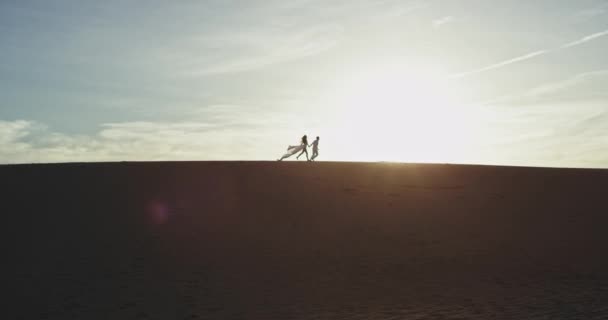 On the large beach side at sunset amazing romantic couple running through the sand in front the camera faraway — Stock Video