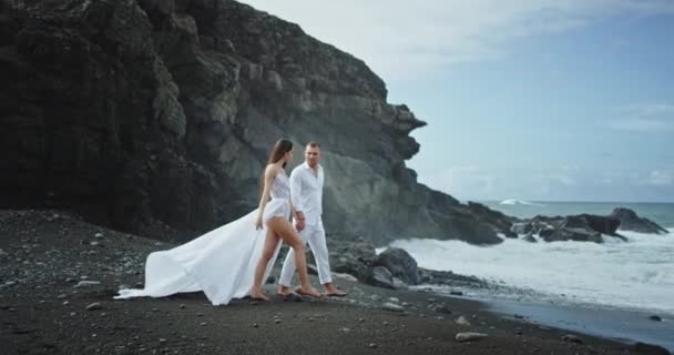 Casal romântico muito atraente tudo em branco eles andando pela praia com areia preta eles são muito felizes apreciando o tempo em vista paisagem incrível — Vídeo de Stock