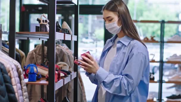 Гарненька леді-клієнт в захисній масці має день покупки, вона бере взуття в руку з підставки і аналізує їх. 4k — стокове відео