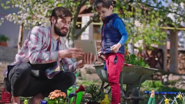 Bell'uomo con il suo piccolo figlio in giardino si divertono a piantare piante papà mostrando al bambino qualcosa sulla tavoletta — Video Stock