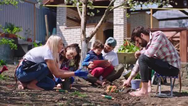 Gran grupo de amigos tienen un gran tiempo juntos en el jardín que plantar algunas plantas y flores en el suelo, excepto la naturaleza papá y su hijo tomar un descanso y jugar un juego en la tableta. Le dispararon. — Vídeos de Stock