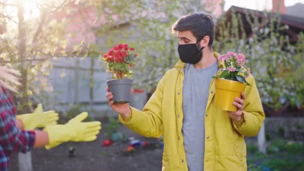 Ung man med en skyddande mask och hans dam med blont hår i karantän i mitten av trädgården de håller blommorna krukor och känner sig glada hemma när du arbetar i trädgården — Stockvideo