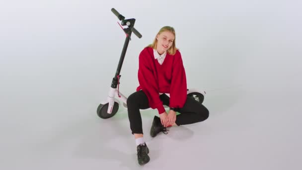 Charismatische blonde dame met een leuke glimlach in de studio met een witte achtergrond zitten naast de electro scooter en recht naar de camera — Stockvideo