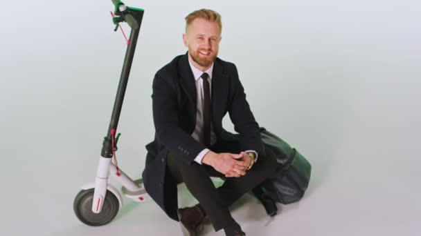 Sullo sfondo con uno sfondo bianco giovane uomo d'affari seduto accanto al suo scooter elettrico con indosso un vestito nero guarda dritto alla fotocamera — Video Stock