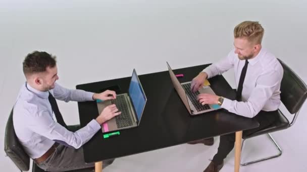 Два бизнесмена в белой студии сидят на своем рабочем месте, используя ноутбуки, на которых они работают, сконцентрировались — стоковое видео