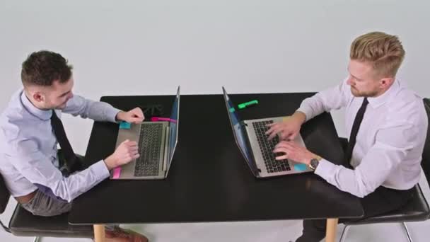 Estudio de fondo blanco dos hombres carismáticos trabajadores de oficina usando dos computadoras portátiles en la misma mesa y escribiendo y discutiendo juntos — Vídeos de Stock