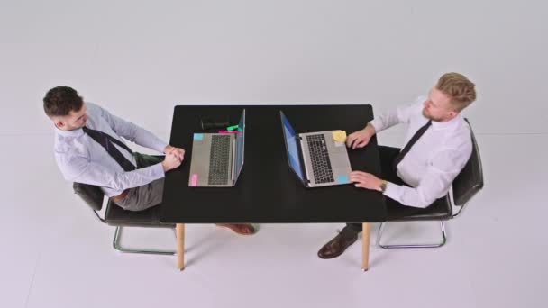 Bien parecido dos trabajadores de oficina sentados en la misma mesa en un gran estudio blanco que utilizan computadoras portátiles para trabajar escribiendo algo — Vídeo de stock