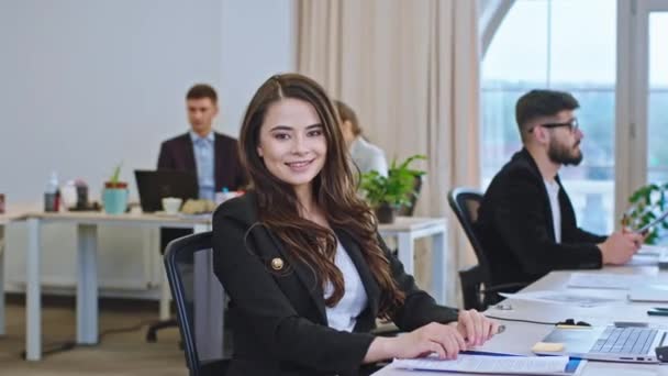 Hermosa señora asistente de oficina en una oficina moderna mientras está sentada en su lugar de trabajo mirando directamente a la cámara y hacer una cara linda — Vídeos de Stock