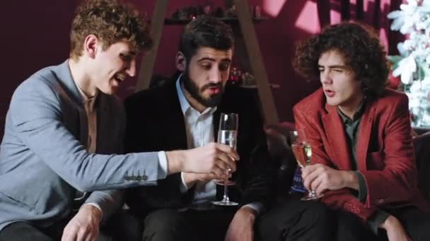 Добре виглядають молоді хлопці на Різдвяній вечірці, сидячи на дивані вони п'ють ігристе вино — стокове відео