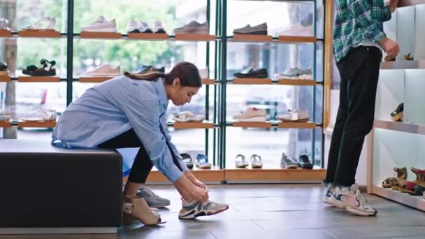 Señora cliente feliz probando algunos zapatos en una zapatería se sienta en el sofá otro tipo cliente mirando a través de los estantes — Vídeos de Stock