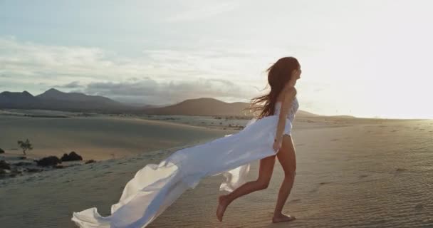 Señora carismática con un largo vestido blanco corriendo hacia su hombre a través de la arena en un lugar increíble al atardecer. 4k — Vídeo de stock