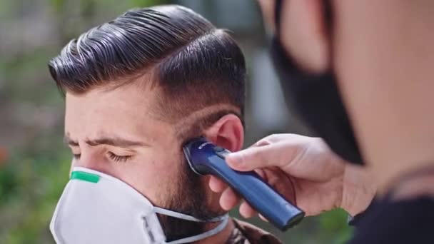 Gros plan en face de l'homme de la caméra ont une coupe de cheveux à la maison dans le jardin dans le salon de coiffure pandémique en utilisant la tondeuse à cheveux qu'ils portent un masque de protection.. 4k — Video