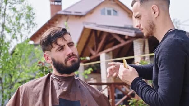 Концентрований чоловік робить свою бороду вдома в саду професійний перукар бере ножиці і вирізає волосся у вигляді бороди — стокове відео
