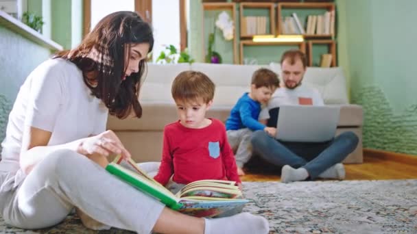 Dobře vypadající matky se svým roztomilým chlapcem čtení příběhu, zatímco sedí na podlaze pozadí táta s druhým dítětem sedí na pohovce a dívá se na něco na notebooku — Stock video