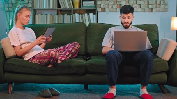 Jong stel thuis in de woonkamer zittend op de sofa dame neem haar elektronische tablet en laat iets zien aan een man heks hebben een laptop op benen — Stockvideo