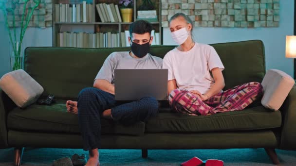Jeune couple en quarantaine à la maison portant un masque de protection qu'ils utilisent un ordinateur portable pour discuter avec leurs amis de la webcam — Video