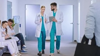 Geniş, modern bir hastane koridorunda iki genç hemşire dijital tablet kullanarak hastanın teşhisini analiz ediyor ve kameranın önünde yürüdüklerini tartışıyorlar.