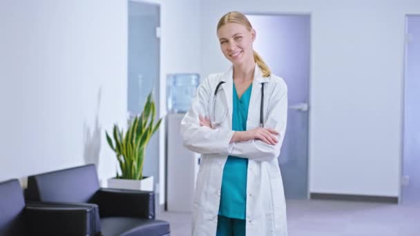 Carismático jovem médico em um hospital moderno no corredor olhando diretamente para a câmera sorrindo grande ela vestindo em seu pescoço o estetoscópio — Vídeo de Stock