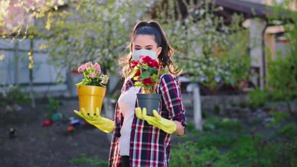 Дома в карантине леди в защитной маске в саду она держит два цветочных горшка и стоит перед камерой в Ковид-19 — стоковое видео