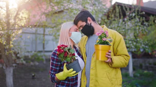 Signora e uomo si sostengono a vicenda nella quarantena di casa si abbracciano mentre tengono in mano due vasi di fiori con indosso la maschera protettiva Covid-19 — Video Stock