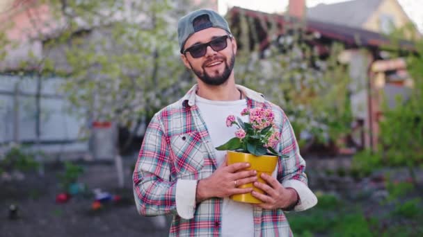 Hombre muy atractivo con una gran sonrisa sosteniendo delante de la maceta de flores de la cámara en el jardín en casa que cuidar después de la naturaleza — Vídeos de Stock