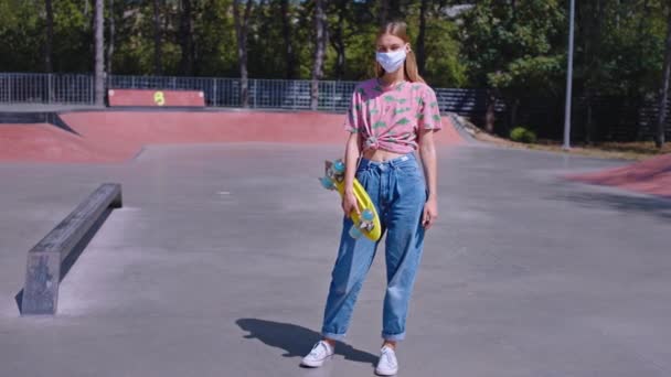 Bonita jovem hipster estilo mulher em um parque de skate urbano na frente da câmera segurando seu skate ela usando máscara protetora para proteger do vírus de COVID-19 — Vídeo de Stock