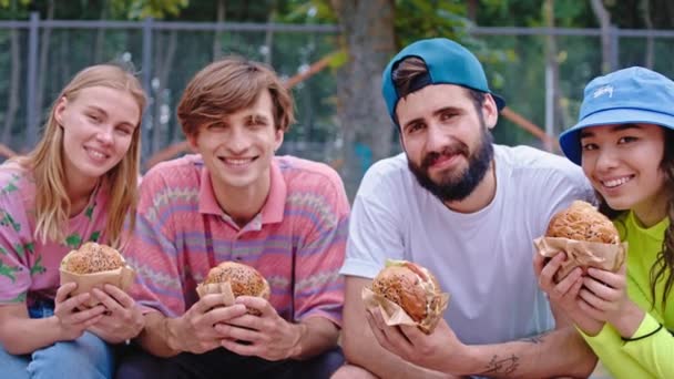 Heureux et souriant grand devant le gros plan ont un temps de pause manger un hamburger après une balade difficile avec skateboard ils assis sur le sol dans un skate park — Video