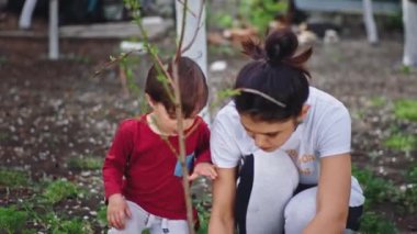 Bahçedeki kameranın önünde oğluyla ilgilenen kadın, ağaçtan sonra bir fırça alıp ağacı beyaz tebeşirle boyuyor.