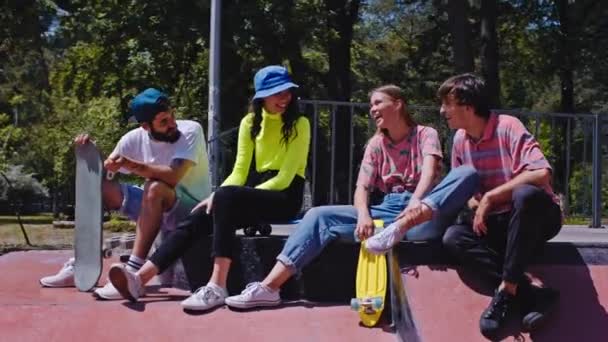 Em um bom dia ensolarado grupo de amigos muito atraente multiétnico aproveitando o tempo em um parque de skate moderno eles sentados no chão e socializar antes de começar a andar de skate. 4k — Vídeo de Stock