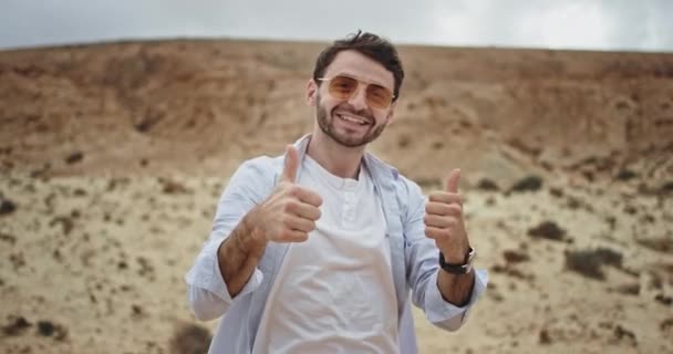Vzrušený turista pohledný muž s velkým úsměvem před kamerou ukazuje velký jako uprostřed úžasné krajiny s kameny a pískem — Stock video