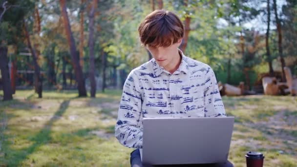 新鮮な空気で勉強するためにノートパソコンを使用して美しい大規模な公園の学生の男で彼はより集中するコーヒーを取る — ストック動画