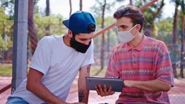 Koncepcja Coronawirus dwóch facetów spotykających się w parku w masce ochronnej zabiera nowoczesny tablet elektroniczny do pracy na świeżym powietrzu nad projektem college 'u — Wideo stockowe