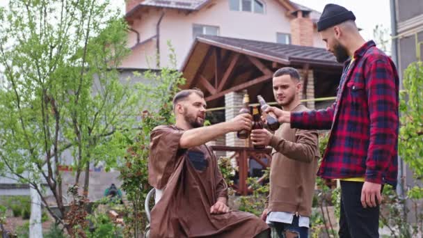 À la maison dans le jardin, trois types charismatiques rencontrant l'un d'eux font une coupe de cheveux à un autre gars pendant qu'ils boivent de la bière — Video