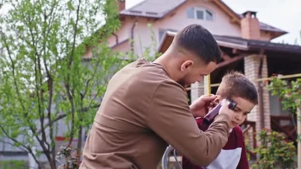En el jardín un hermano mayor hacer el corte de pelo a su hermano pequeño que el uso de la cortadora de pelo que tienen un buen humor — Vídeos de Stock