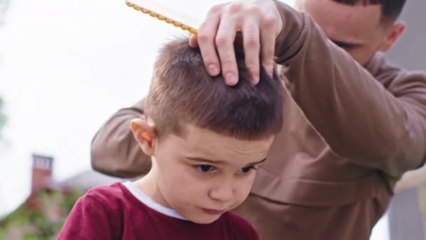 Carismatico piccolo ragazzo primo piano per la fotocamera in mezzo al giardino seduto sulla sedia e in attesa di finire il taglio di capelli l'uomo barbiere — Video Stock