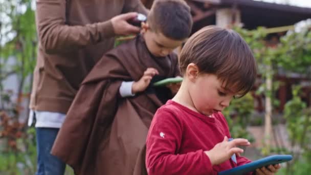 Zwei charismatische kleine Jungen, einer sitzt auf dem Smartphone und wartet, während sein Bruder draußen im Garten seinen Haarschnitt beendet. — Stockvideo