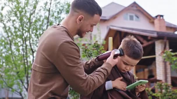 Pequeño niño divertido concentrado jugando en el teléfono inteligente, mientras que el peluquero profesional terminará el corte de pelo con la cortadora de pelo — Vídeo de stock