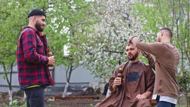 Na frente da câmera homem carismático no jardim ter uma conversa com seus amigos que bebem cerveja, enquanto ele tem um corte de cabelo com um barbeiro profissional — Vídeo de Stock