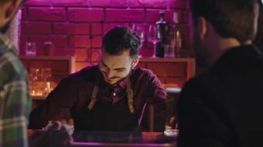 Bira barında iyi görünümlü Bart çalışırken büyük bir gülümsemeyle bar masasını temizlerken diğer müşteriler bar masasında iyi vakit geçiriyorlar.